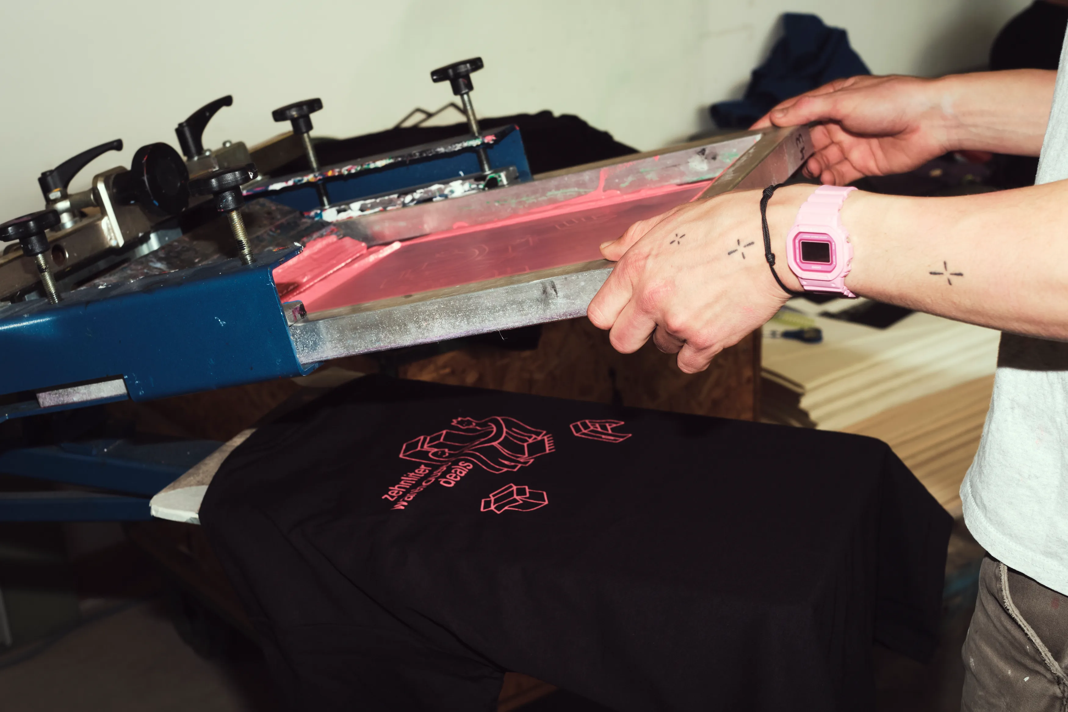 Hands pulling a squeegee across a screen to print red ink on a black shirt.